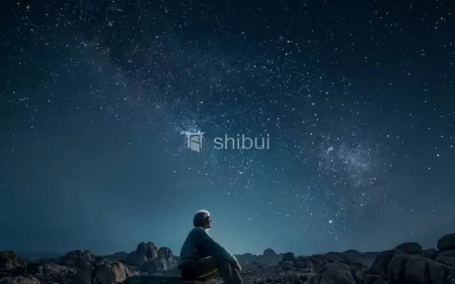 Um homem sentado em rochas observa o céu noturno repleto de estrelas e a Via Láctea.