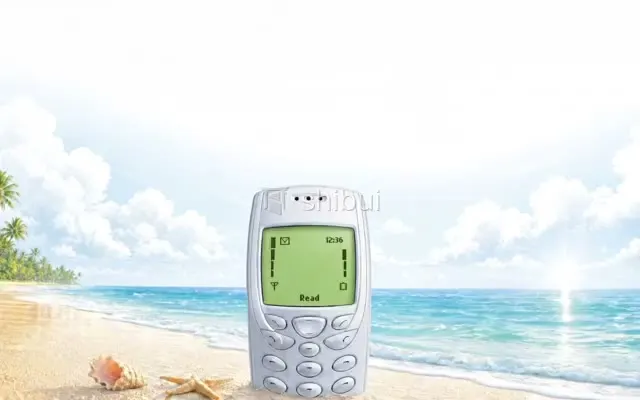 Um celular antigo na areia de uma praia tropical com o mar azul ao fundo e céu com nuvens.