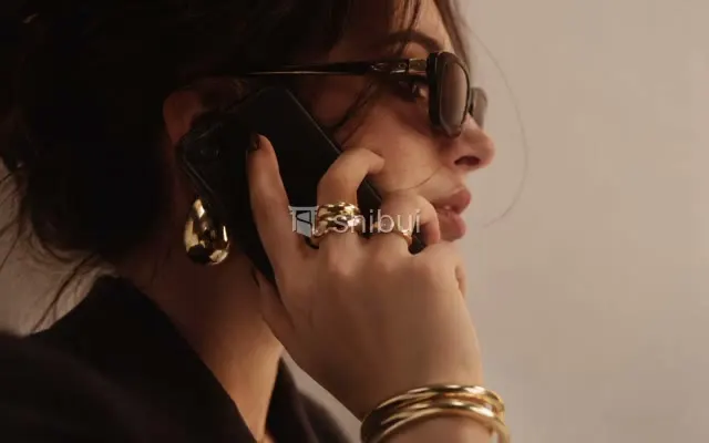Mulher de óculos escuros fala ao celular, usando anéis e brincos dourados chamativos.
