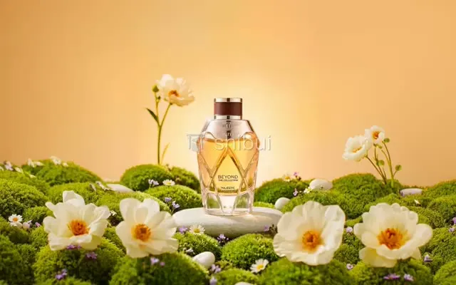 Um frasco de perfume dourado repousa sobre pedras em um jardim de musgo com flores brancas e um fundo alaranjado.