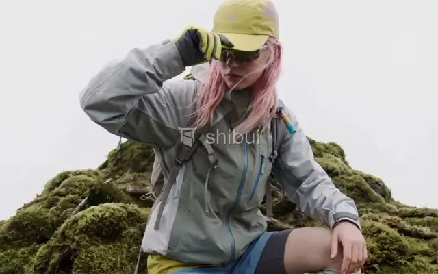 Uma pessoa com cabelo rosa e roupa de montanha sentada em uma colina coberta de musgo verde.