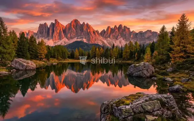 Montanhas refletidas em um lago calmo com céu alaranjado e árvores verdes e amarelas ao redor.