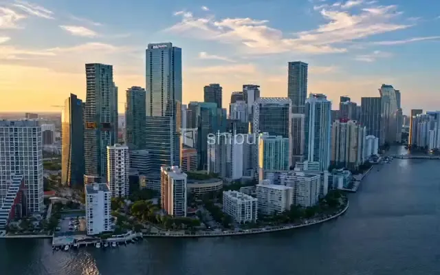 Uma vista aérea de arranha-céus modernos refletindo a luz dourada do pôr do sol sobre a água.