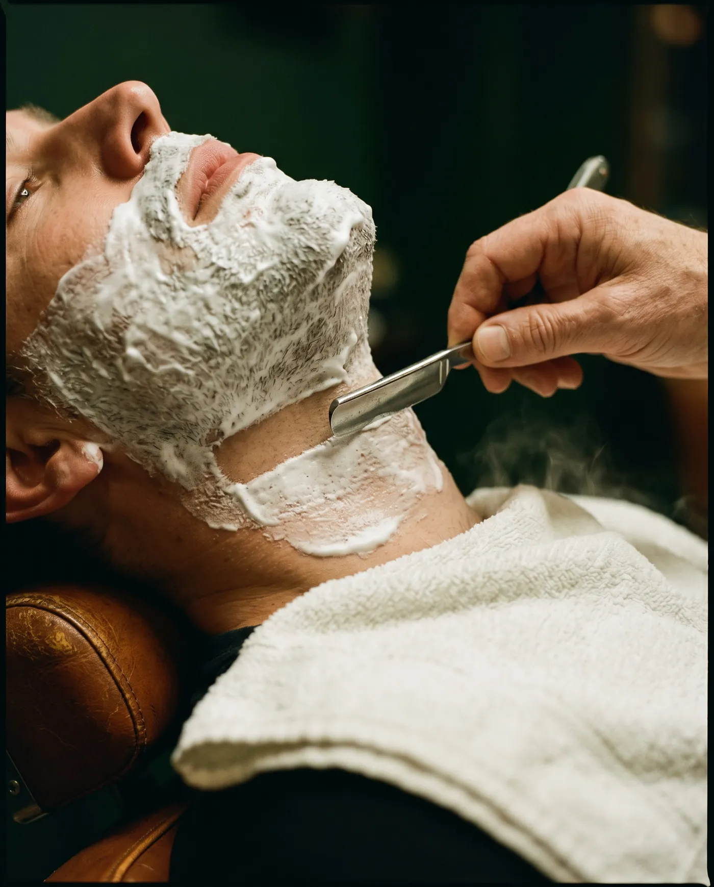 Barbeiro fazendo a barba de um cliente com navalha, espuma branca e toalha quente